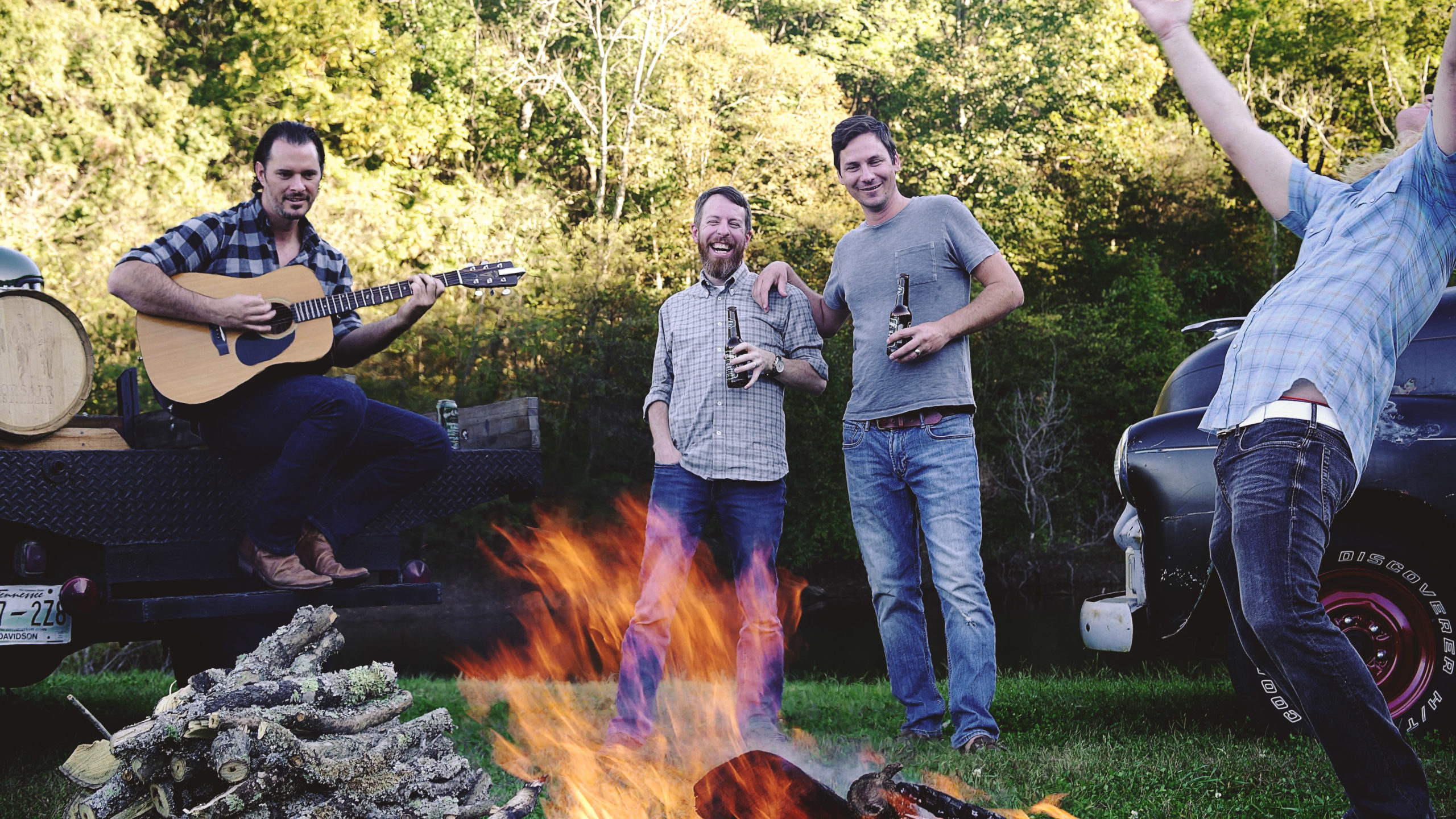 4 men drinking beer and playing guitar around a fire